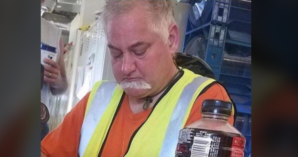 A middle-aged man with gray hair and a mustache is sitting and appears to be dozing off. He is wearing an orange shirt and a high-visibility safety vest. In the foreground, there is a bottle of soda on the table and some industrial equipment in the background.