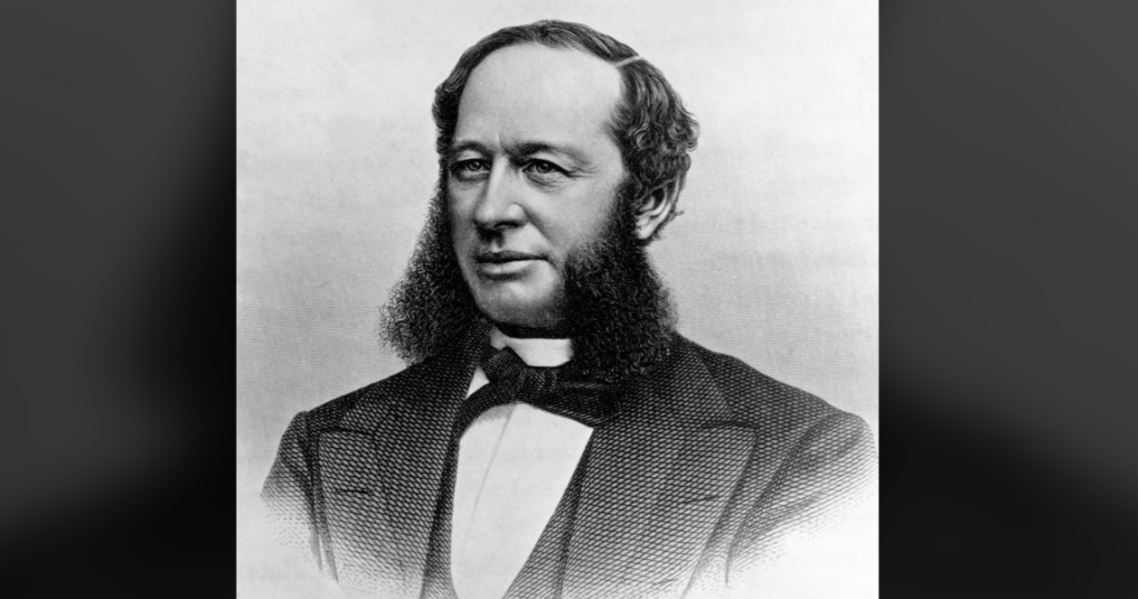 A black and white portrait of a man with a suit and tie. He has thick, prominent sideburns extending down his cheeks and a slight smile. The background is plain, emphasizing his formal attire and distinctive facial hair.