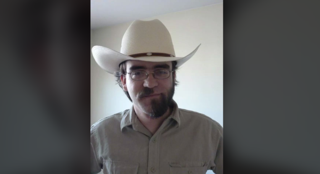 A person with glasses, a beard, and mustache is wearing a beige cowboy hat and a matching shirt with front pockets. The person is indoors, standing against a plain light-colored wall.