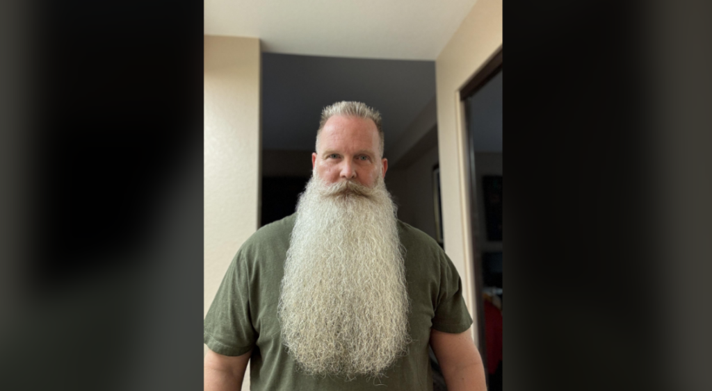 A man with a long, flowing white beard and short, styled hair stands facing the camera. He is wearing a green t-shirt and is set against a background of neutral-colored walls and doorways.