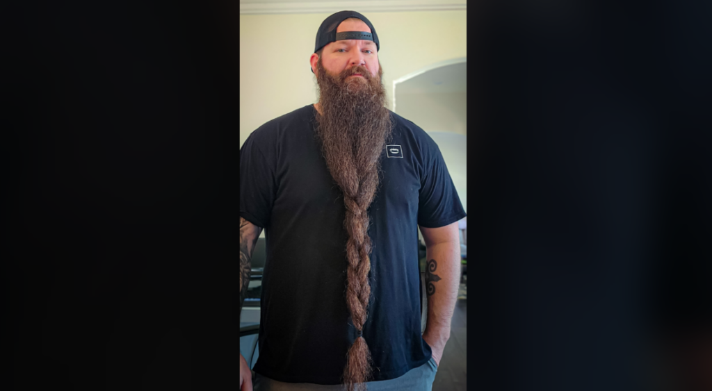 A man with a long, braided beard stands indoors. He is wearing a black T-shirt, a black baseball cap worn backwards, and has tattoos on his arms. The beard extends well past his waist. He appears to be in a home environment.