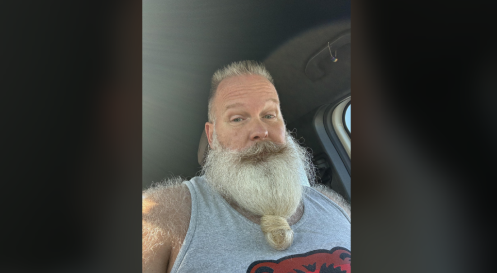 A bearded man with a white and thick beard tied in a knot at the end is sitting in a car. He is wearing a sleeveless shirt featuring a red and black bear graphic. Sunlight filters into the vehicle, highlighting his face and shoulders.