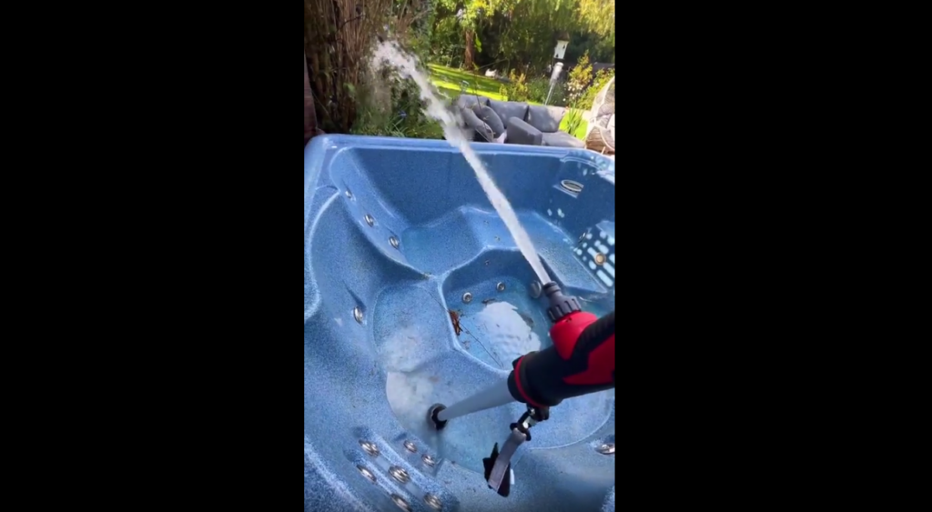 A person is cleaning an empty blue hot tub outdoors with a high-pressure spray nozzle attached to a hose. Water is being sprayed onto the hot tub's surface to wash away dirt and debris. Trees and greenery are visible in the background.