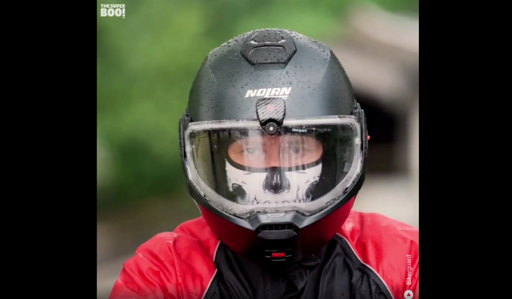 A person wearing a black Nolan motorcycle helmet with a transparent visor that shows a skull design covering their face. They are dressed in a red and black jacket, with water droplets visible on the helmet. The background is blurred with green and gray hues.