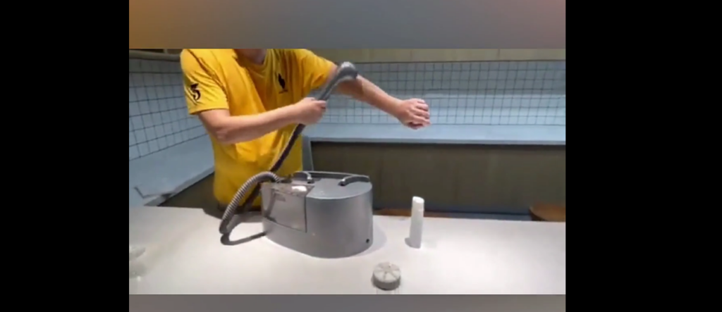 A person wearing a yellow shirt is using a handheld spray nozzle to spray their arm. The nozzle is connected to a grey machine placed on a kitchen counter. A small bottle and a round object are also on the counter nearby. The background features tiled walls.