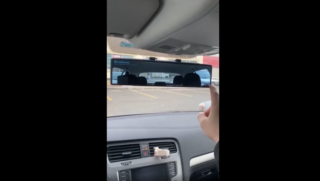 A person inside a car points at the rearview mirror which displays the parking lot and a building behind the car. The dashboard features a car audio system and an air vent, with the windshield displaying part of the car's interior roof and mirror mount.