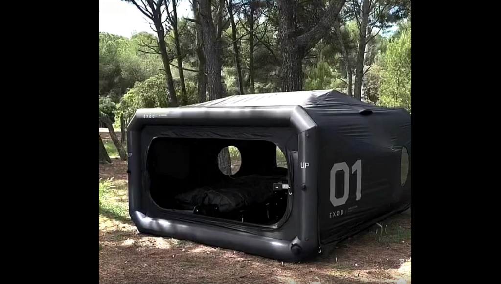 An inflatable black tent labeled “01” is set up in a wooded area. The front opening reveals an interior with a bed. Tall trees and greenery surround the tent, creating a serene camping environment.