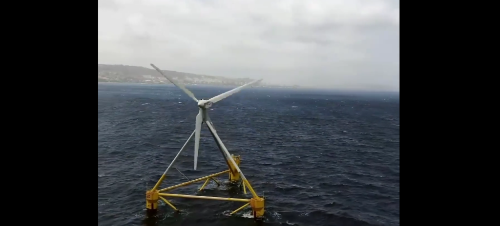 A large wind turbine stands on a floating platform in the ocean, with choppy waves surrounding it. The platform is secured by yellow triangular structures. In the background, a distant coastline is visible under a cloudy sky.