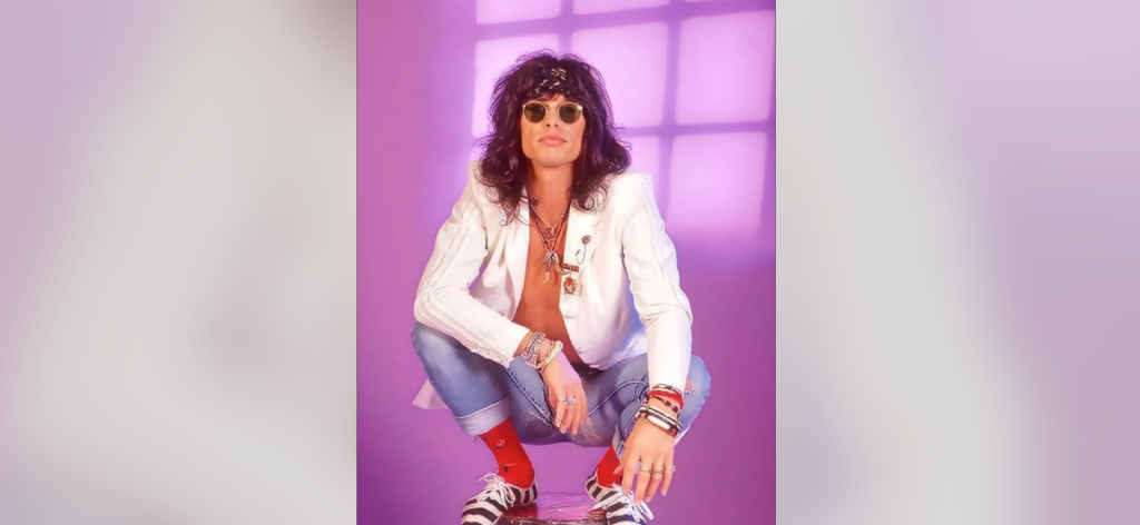 A person with long hair and sunglasses squats against a purple background. They wear a white jacket, blue jeans, red socks, and black-and-white striped shoes. The person also has several bracelets and a bandana on their head, exuding a casual, rebellious vibe.