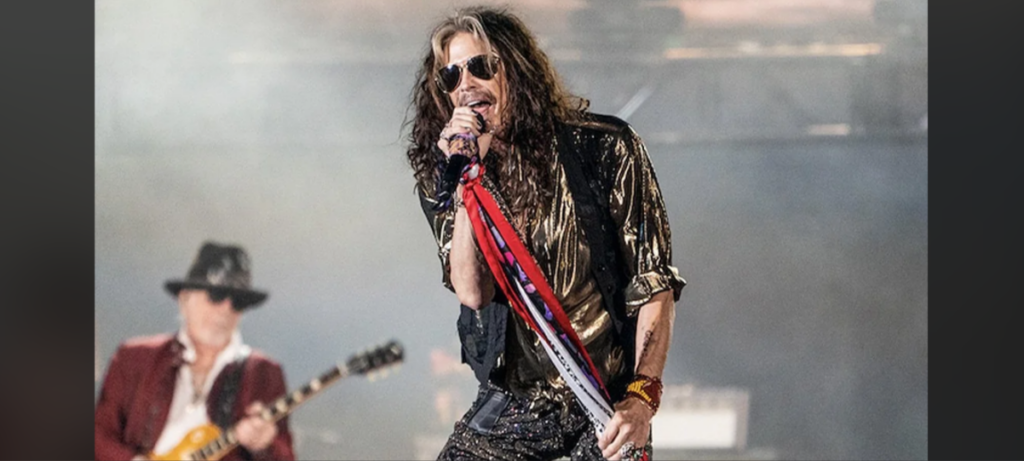 A male rock singer with long hair, wearing sunglasses and a metallic shirt, holds a microphone stand draped with scarves and leans forward singing. A guitarist in a fedora and sunglasses plays in the background. Smoke rises, enhancing the stage lighting.