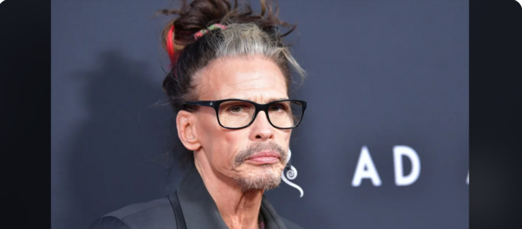 A man with long hair tied up in a messy bun, wearing black-framed glasses and a dark shirt, is posing against a dark backdrop. His hair is mixed with grey and he has a serious expression on his face.