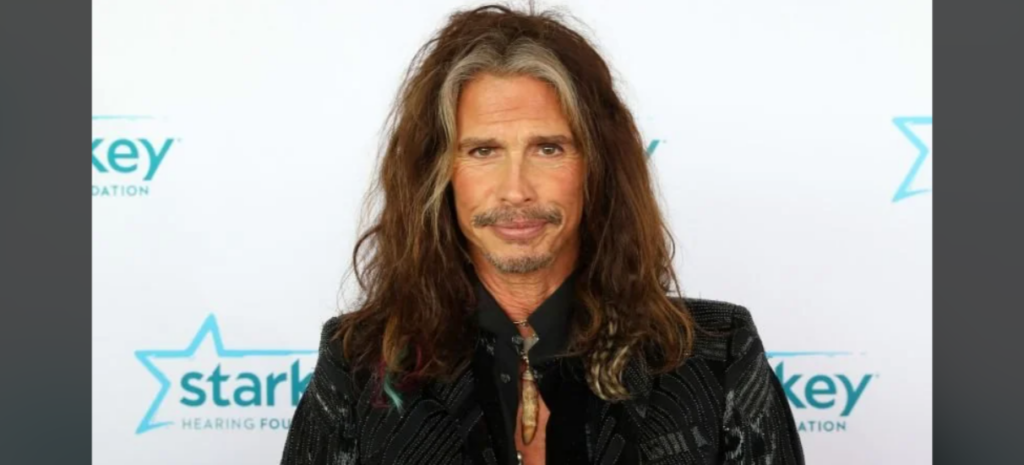 A person with long, wavy hair wearing a black patterned jacket is standing in front of a backdrop with the Starkey Hearing Foundation logo. They have a slight smile and a necklace with a pendant.