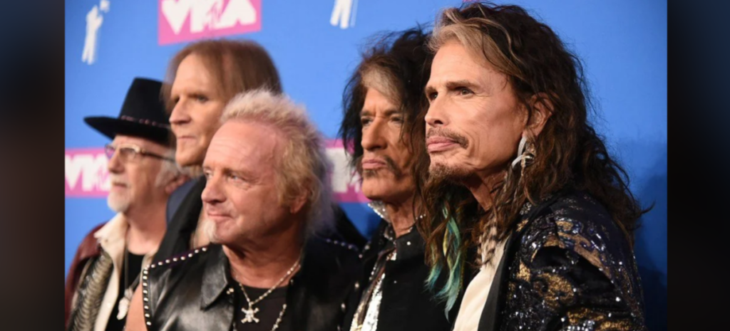 A group of six men stand together against a blue backdrop with white and pink logos. They are wearing rock-style attire, including leather jackets, chains, and hats. The men have long hair, and some have facial hair, showcasing a rock band vibe.