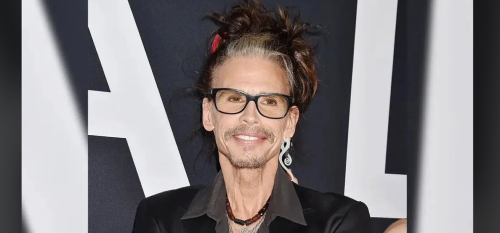 A person with long, styled hair, black glasses, and a mustache and goatee poses for a photo against a dark background with white lettering. They are wearing a black jacket and a red necklace. The individual looks towards the camera with a slight smile.