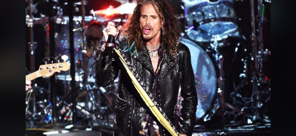 A long-haired person wearing a black leather jacket is singing passionately into a microphone. They are holding a microphone stand wrapped with yellow fabric. Drums and other musical instruments are visible in the background, suggesting a live rock performance.