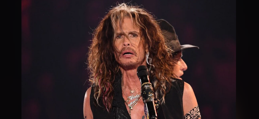 A person with long, wavy hair, wearing a sleeveless top and various necklaces, is seen singing into a microphone on stage. Another individual in a hat is in the background. The stage lighting is dim with a dark, blurred backdrop.