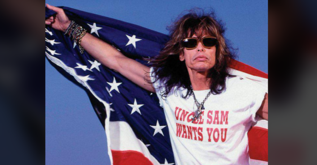 A person with long hair wearing sunglasses holds out an American flag behind them. They wear a white T-shirt with the text "Uncle Sam Wants You." The background is a clear blue sky.