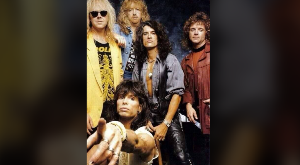 A group of five men pose for a photo against a dark background. The man in the front extends his hand towards the camera. They have long hairstyles and wear a mix of casual and leather clothing, including jackets and sunglasses. The mood is intense and expressive.