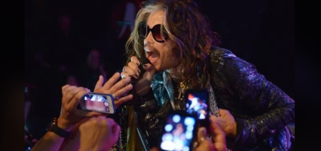 A rock musician with wavy hair and wearing sunglasses passionately sings into a microphone while leaning towards an excited crowd. Fans' hands and smartphones are seen reaching towards the singer, capturing the moment. The background is dark, highlighting the musician.