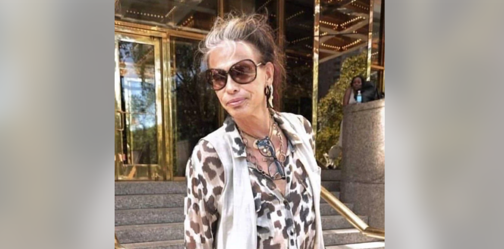 A woman with gray and dark hair, wearing large sunglasses, a leopard print blouse, and a beige vest is standing outside a building with glass doors and a gold trim. She is accessorized with necklaces and earrings. The scene includes steps and a railing.