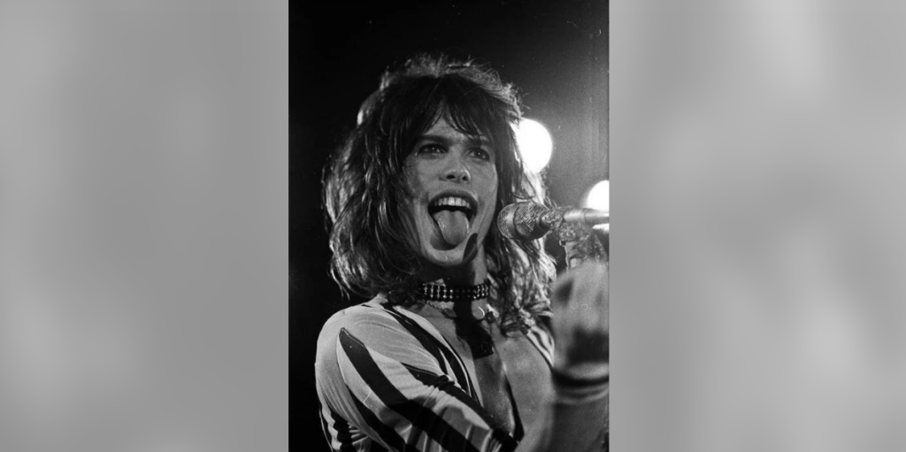 A black-and-white photo of a long-haired person on stage, wearing a striped shirt and a choker necklace. They are singing passionately into a microphone, with their mouth open and tongue extended. Bright stage lights are visible in the background.