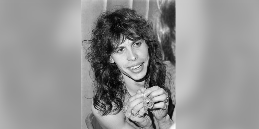 Black and white photo of a person with long wavy hair smiling, holding a small object in their hands. They are visible from the shoulders up and dressed in a sleeveless top. The background is plain and slightly blurred.