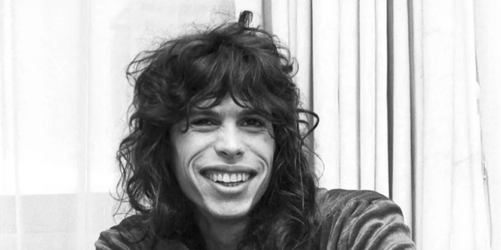 A person with long, curly hair is smiling widely while looking at the camera. They are indoors in front of light-colored curtains. The image is in black and white.