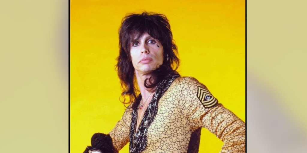 A person with shoulder-length dark hair stands against a bright yellow background, wearing a gold-patterned jacket with black accents and a decorative insignia on the sleeve. They have an intense expression and are looking directly at the camera.