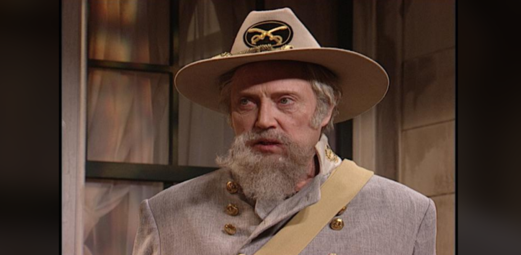 A man with a gray beard and mustache wears a gray Confederate uniform with gold buttons and a wide-brimmed hat featuring an emblem. He stands indoors near a window with beige curtains.