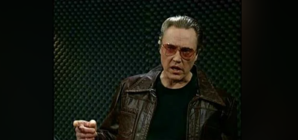 A person wearing sunglasses and a brown leather jacket is standing against a dark, textured background. They appear to be speaking or singing, with one hand raised slightly in a gesturing motion.