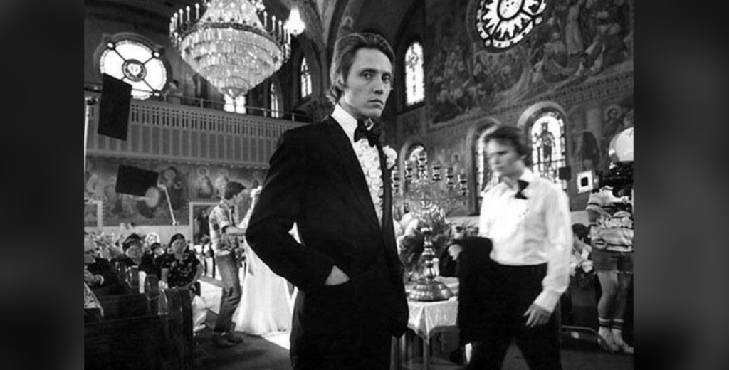 A black-and-white photo of a wedding scene in an ornate church. The foreground shows a man in a tuxedo looking at the camera, with a boutonniere pinned to his jacket. Several people, including another man in a tuxedo, are visible in the background.