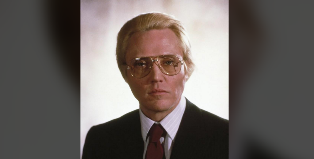 A person with light-colored, slicked-back hair, wearing large glasses and a dark suit with a white shirt and red tie, poses against a neutral background. Their expression is serious.