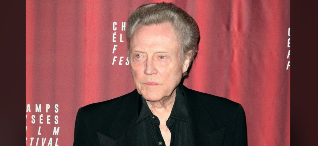 A man with combed back grey hair wearing a black suit and shirt stands in front of a red backdrop with white text at the Champs-Élysées Film Festival.