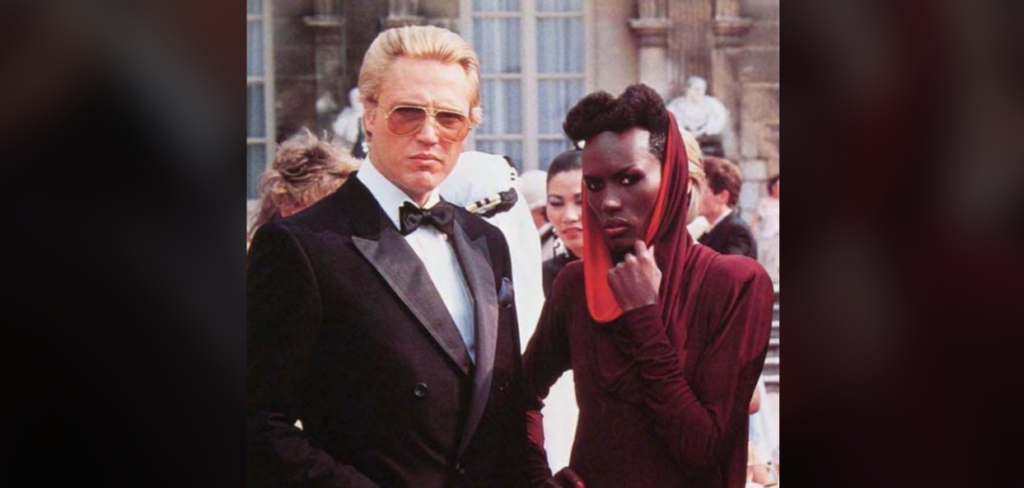 A person wearing a black tuxedo with sunglasses stands next to another person dressed in a deep red outfit with a matching headpiece. The background features an ornate building facade with statues, suggesting a formal event or gathering.