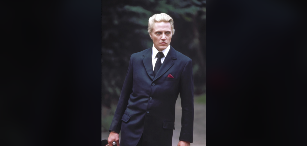 A person with slicked-back blond hair is dressed in a dark suit with a white shirt and black tie, holding a briefcase. The background is blurred, featuring greenery. The person is standing in a confident pose, looking off to the side.