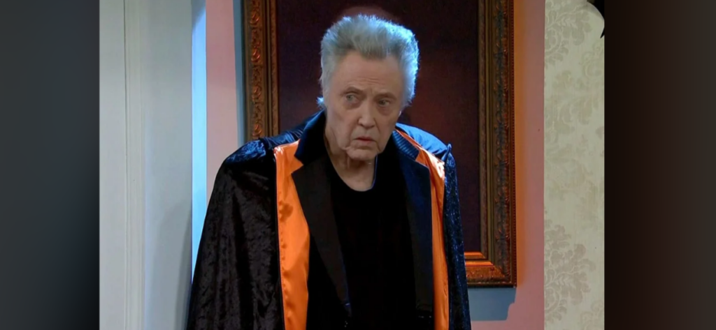 An older man with gray hair stands indoors, looking at the camera with a serious expression. He wears a dark cloak with an orange lining over a black outfit. The background consists of a framed painting and a partial view of wallpapered and painted walls.