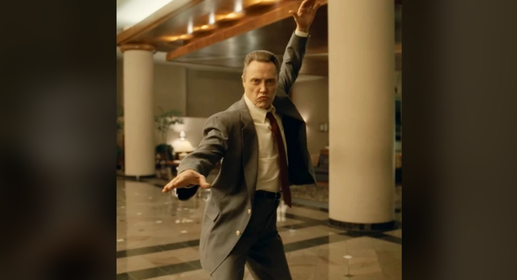 A man in a gray suit is energetically dancing in what appears to be a hotel lobby. He has one arm raised above his head and the other extended sideways. His expression is serious and focused, and the lobby is decorated with columns and warm lighting.