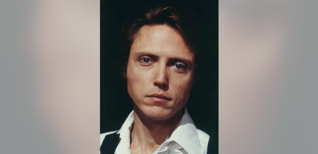 A close-up portrait of a person with a serious expression, short brown hair, and blue eyes, wearing a white shirt and a dark vest. The background is dark, highlighting the person's face.