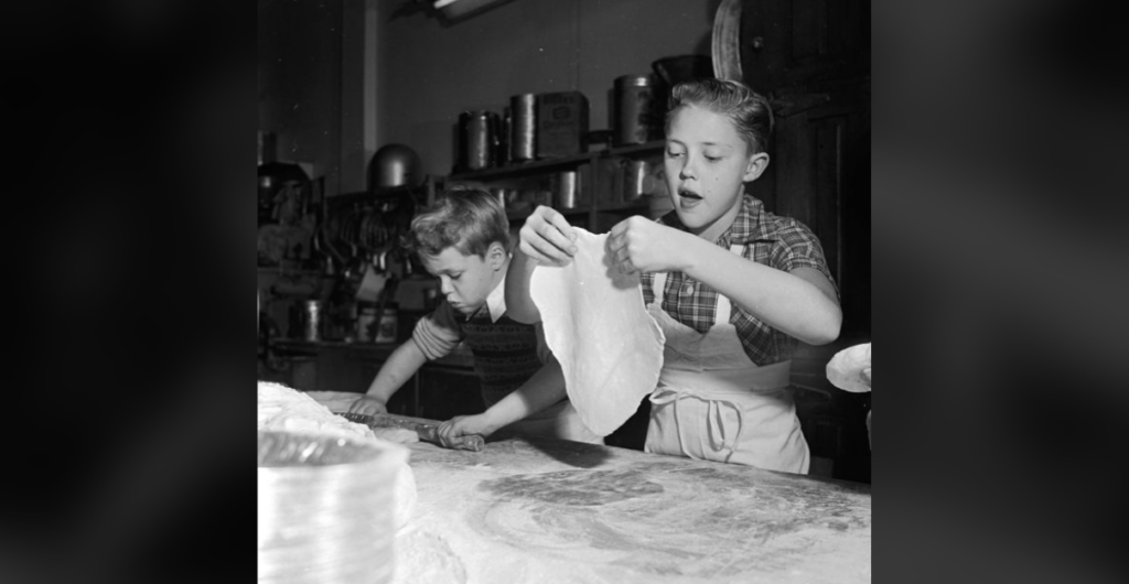 Two boys, both wearing aprons, are engrossed in cooking. The boy on the left is rolling dough with a rolling pin, while the boy on the right is stretching a sheet of dough with his hands. They are in a kitchen with various utensils and containers in the background.