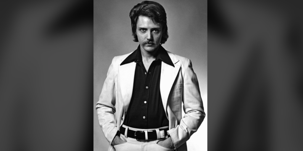 A black and white photograph shows a person with medium-length hair and a mustache. They are wearing a light-colored blazer over a dark shirt with wide collars. They have their hands in the pockets of light-colored pants, and the background is plain.