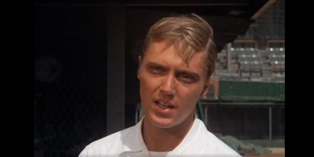 A young man with blonde hair is standing in front of an empty stadium. He is wearing a white shirt and looking slightly past the camera with a neutral expression. The sunlight highlights his features, and the background shows empty bleachers and part of the field.