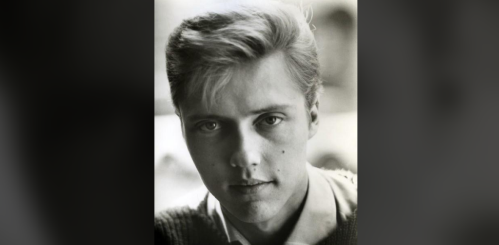 A black-and-white portrait of a young man with short, neatly-styled hair. He gazes directly into the camera with a neutral expression. He is wearing a collared shirt and a sweater. The background is blurred, drawing focus to his face.