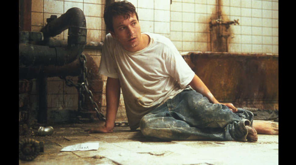 A person wearing a dirty white t-shirt and jeans is sitting on the grimy tiled floor of a dilapidated room. The surroundings are dirty with rusted pipes and stained walls. Papers and other debris are scattered around. The person appears to be in distress.