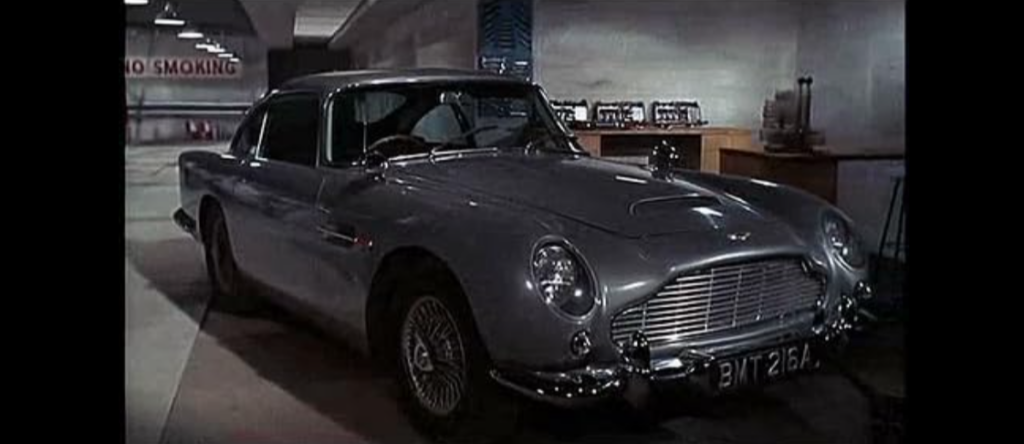 A classic silver Aston Martin DB5 is parked indoors in a well-lit garage with no smoking signs visible. The car features sleek lines, wire wheels, and a prominent front grille. The license plate reads "BMT 216A.