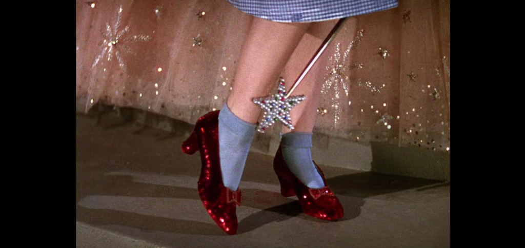 A close-up of a person wearing sparkling red shoes with blue socks. The person is holding a star-tipped wand, and part of a pink, glittery skirt is visible in the background.