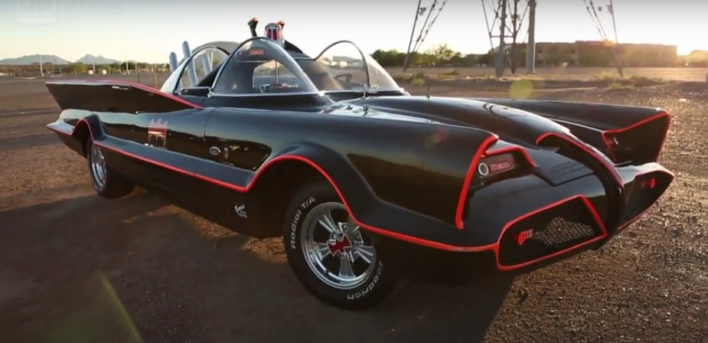 A classic black car with red accents, resembling the iconic Batmobile from the 1960s Batman TV series, is parked on a deserted road. The car features a bubble windshield, bat-shaped motifs, and sleek, aerodynamic design with prominent rear fins.