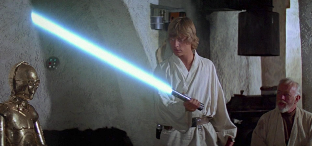 A young man holding a blue lightsaber stands between a golden humanoid robot and an elderly man with a gray beard. The scene appears to be inside a rustic, dimly lit room. The young man looks at the lightsaber in amazement, while the others observe calmly.