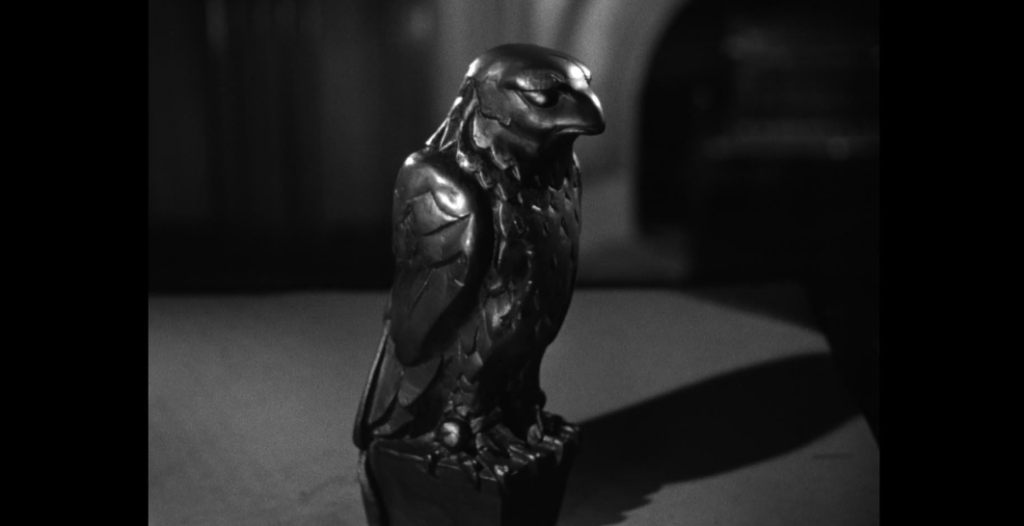 Black-and-white photo of a statuette depicting a bird of prey, likely a falcon, with detailed feathers and a stern expression. The statue rests on a small rectangular base, placed on a flat surface with a blurred background.