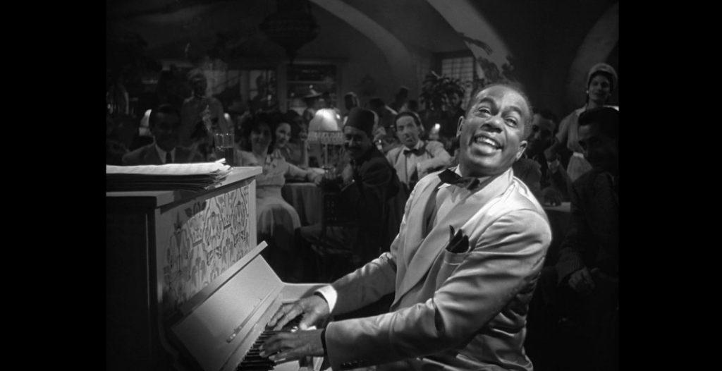 A man smiles broadly while playing a piano in a lively, dimly-lit club. He wears a light-colored suit with a dark bow tie, surrounded by an audience. A shadowed, arching ceiling and a mix of seated and standing people add to the rich atmosphere of the venue.