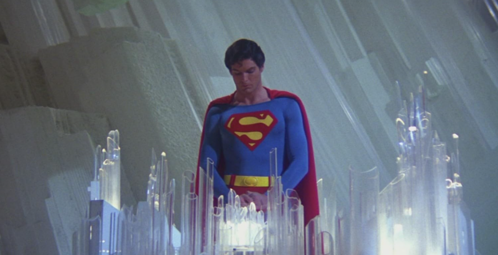 A person dressed in a blue suit with a red cape and an 'S' emblem on the chest stands solemnly before a display of crystal-like structures. The background features a futuristic, crystalline environment.